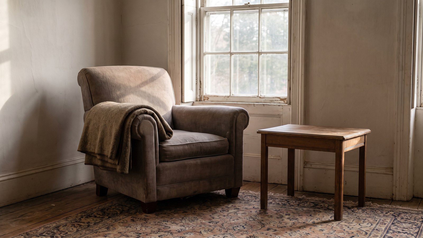 A single upholstered armchair beside a tall window with a folded wool throw over the arm and a bare side table