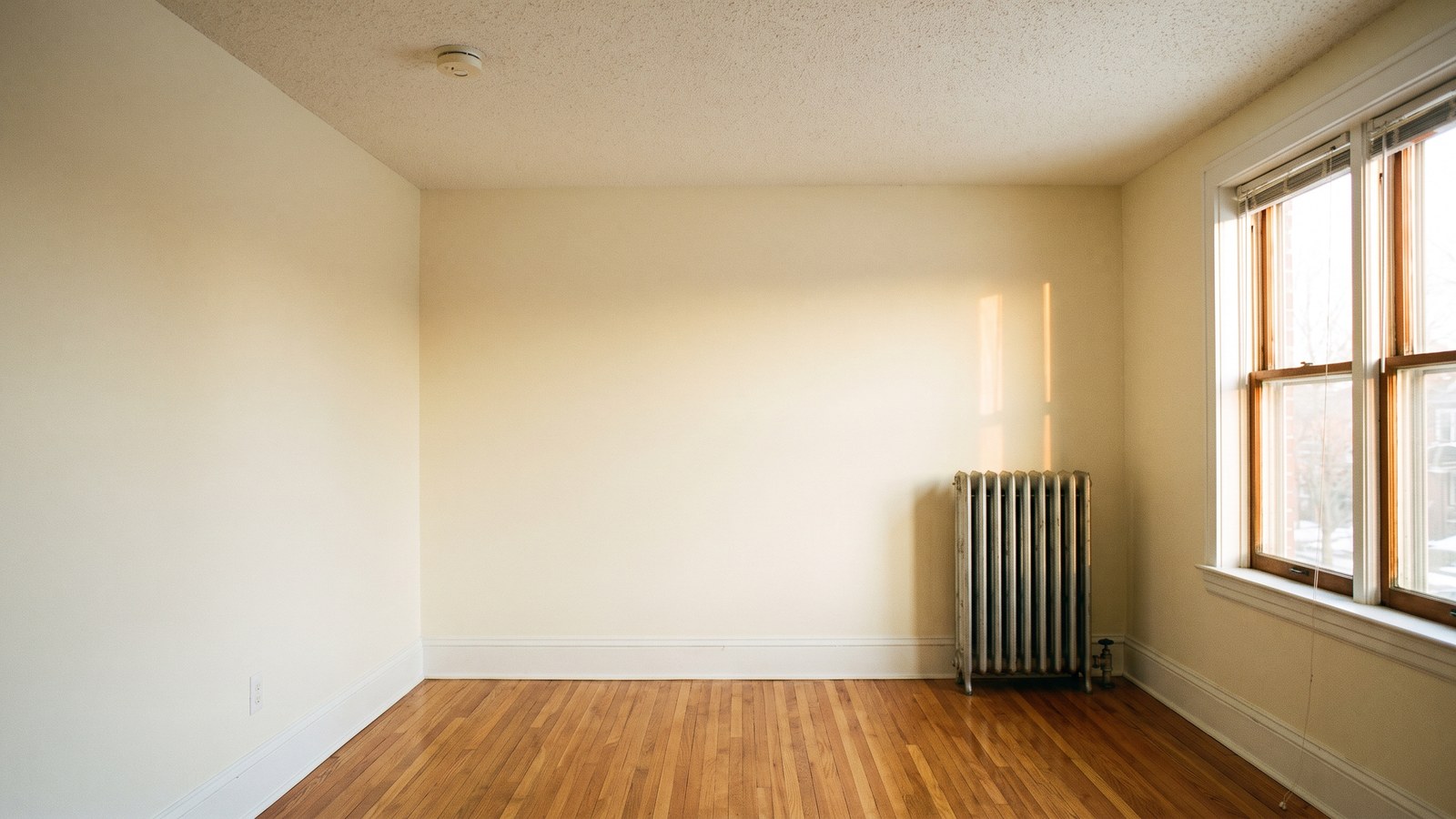 A clean, empty apartment living room in an older house with freshly painted walls, wood floors and an old radiator beside a sunny window