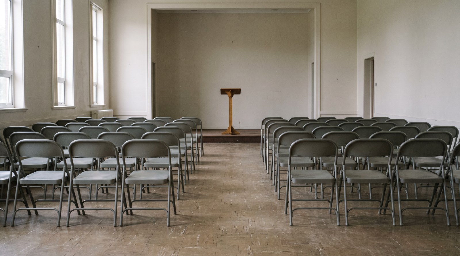 Rows of empty grey folding chairs in a plain community hall facing a simple wooden lectern on a low platform