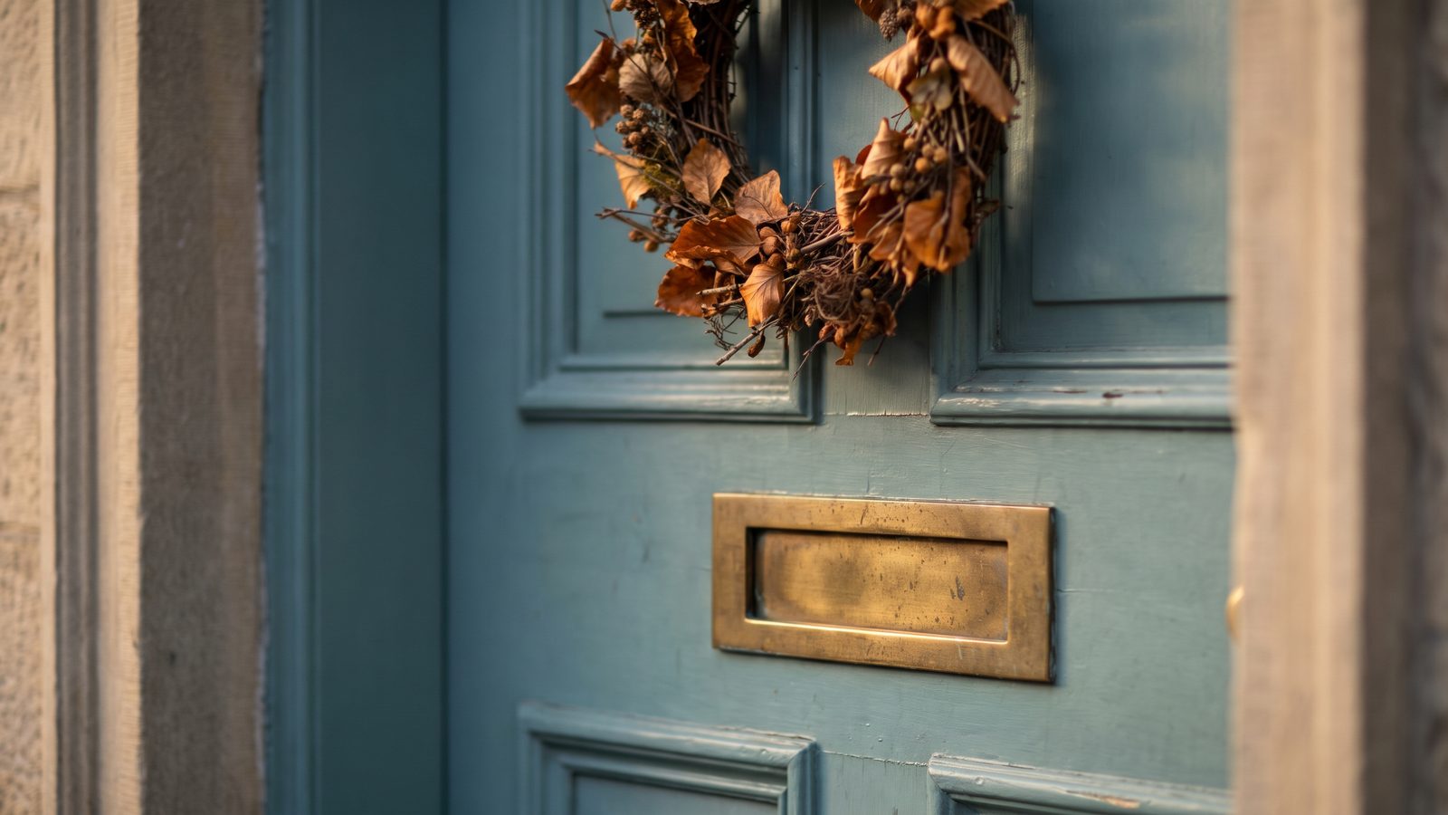 A painted front door with a brass mail slot and a wreath of dried autumn leaves in soft side light
