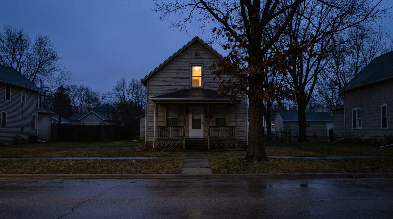 A modest two-storey clapboard house on an ordinary street at dusk, one upstairs window lit from inside