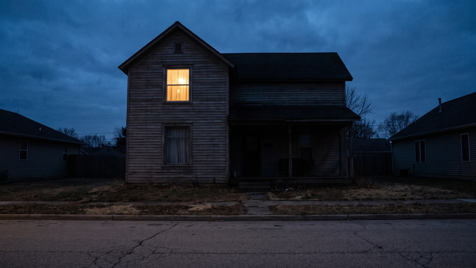 The exterior of a modest older house at dusk with a single upstairs window softly lit from within and the rest of the house dark