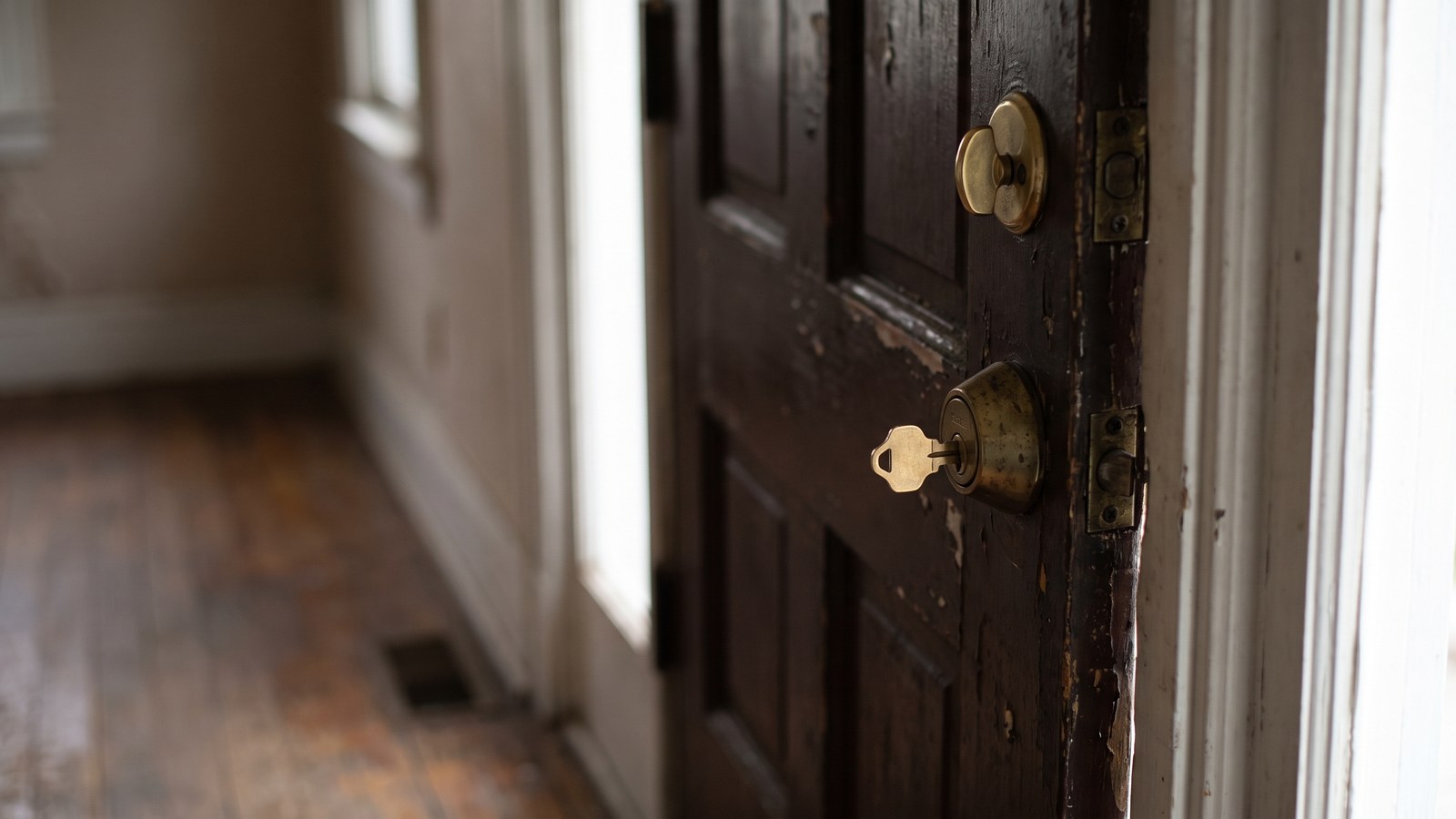 A brass key left in the inside keyhole of a worn wooden front door, seen from a dim hallway