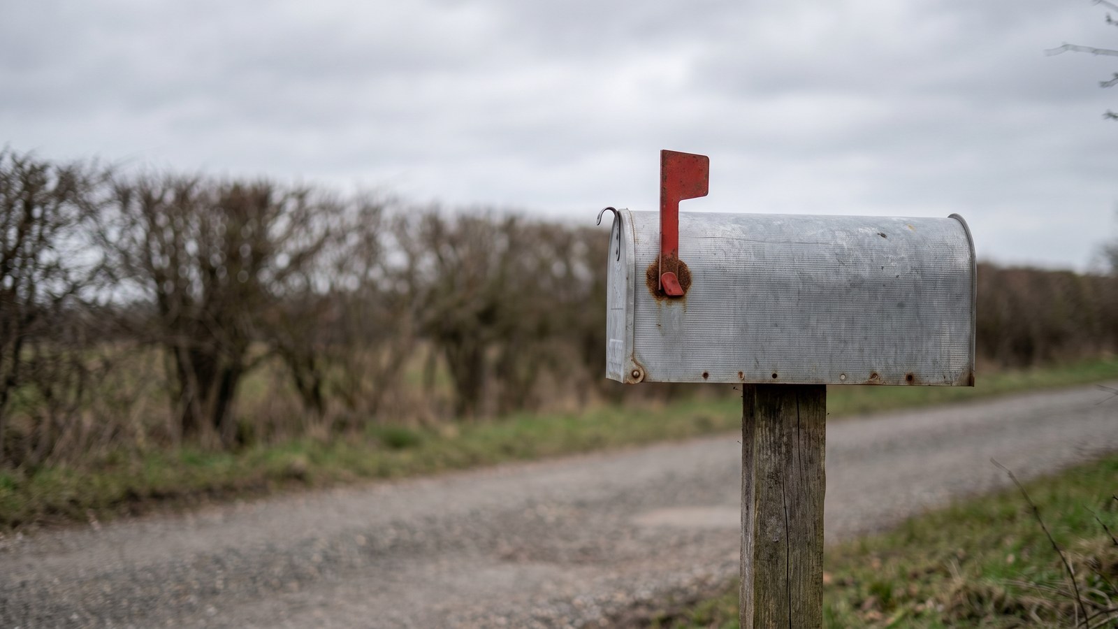 A weathered metal rural mailbox on a wooden post at the end of a gravel driveway with its red flag raised