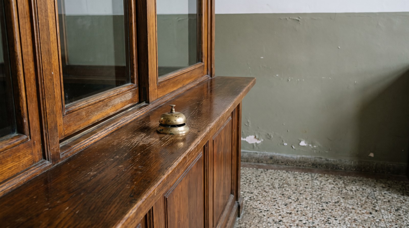 An old varnished wooden public office counter with a closed sliding glass window above it and a tarnished brass call bell