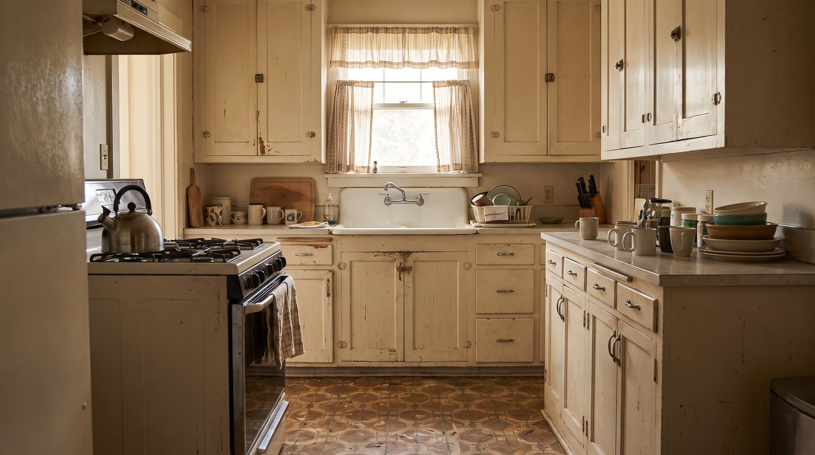 A well-worn older kitchen with painted wooden cabinets from decades ago, a stovetop kettle and a patterned linoleum floor