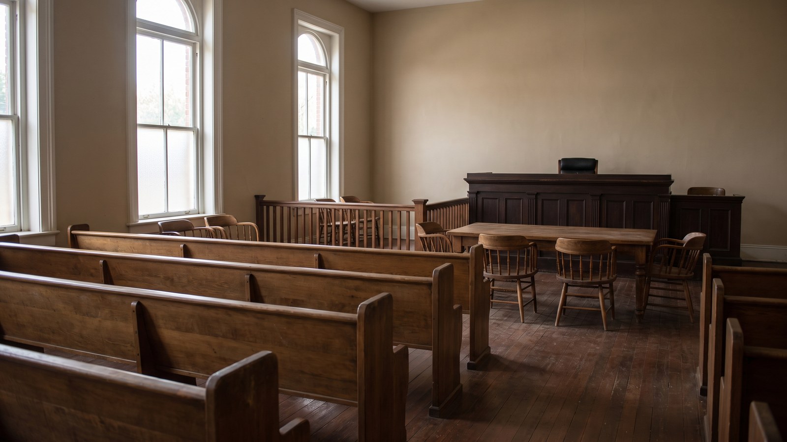 An empty county courthouse hearing room with plain wooden bench seating and a low rail