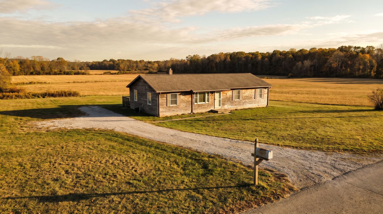 A modest single-story ranch house on a large rural lot with a gravel driveway, open fields and a tree line behind it in late October light