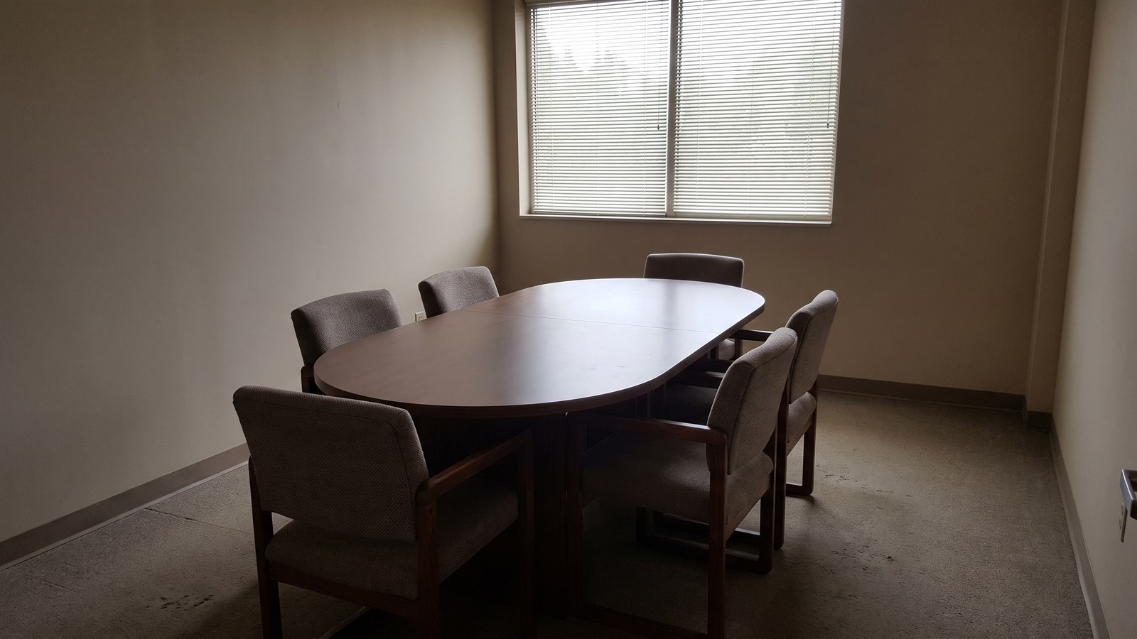 An empty small conference room with a plain oval wooden table, six chairs pushed in and a window with slatted blinds letting in daylight