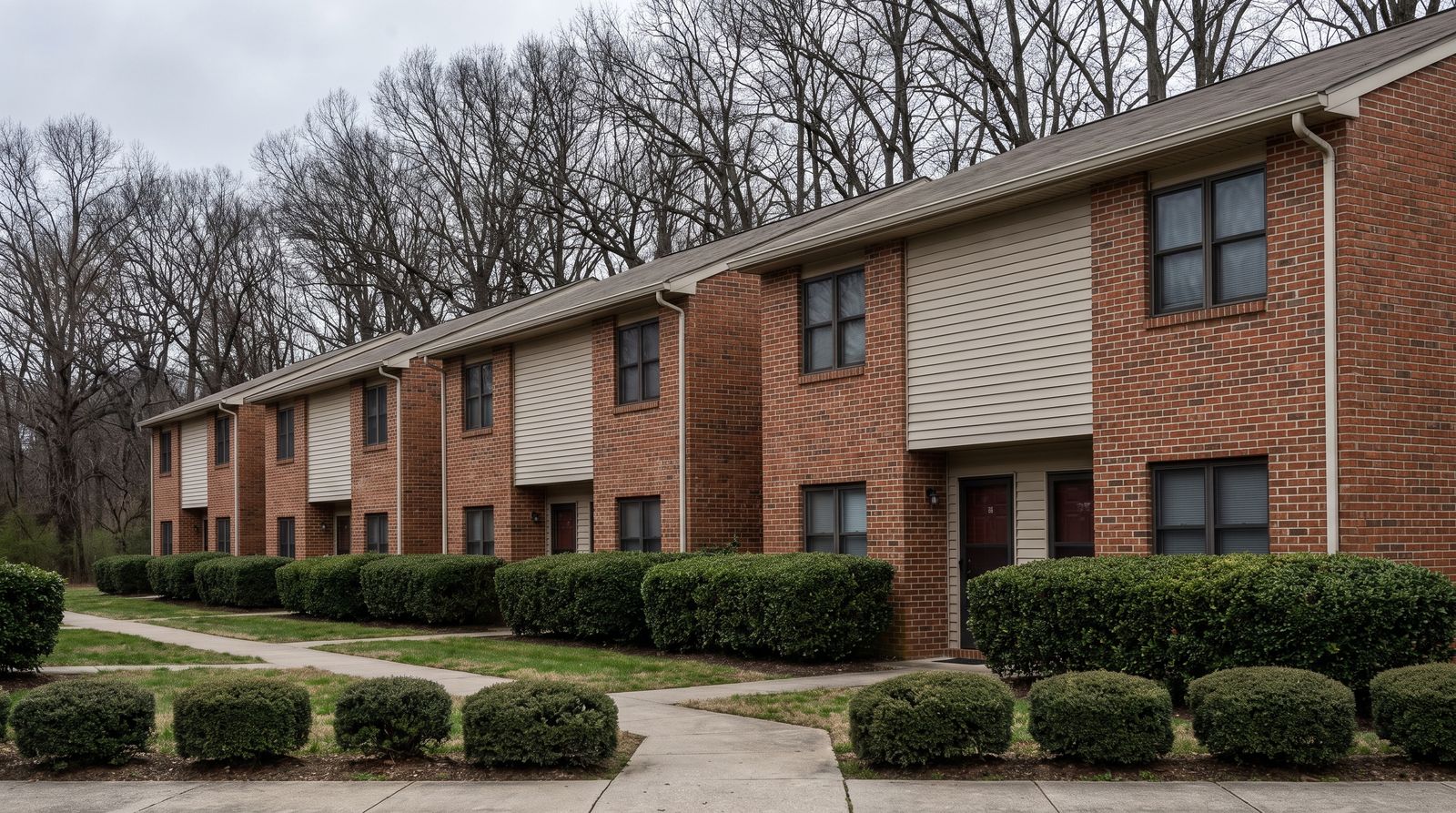 A row of modest two-storey townhouse condominium units with uniform facades, shared clipped landscaping and a communal paved walkway