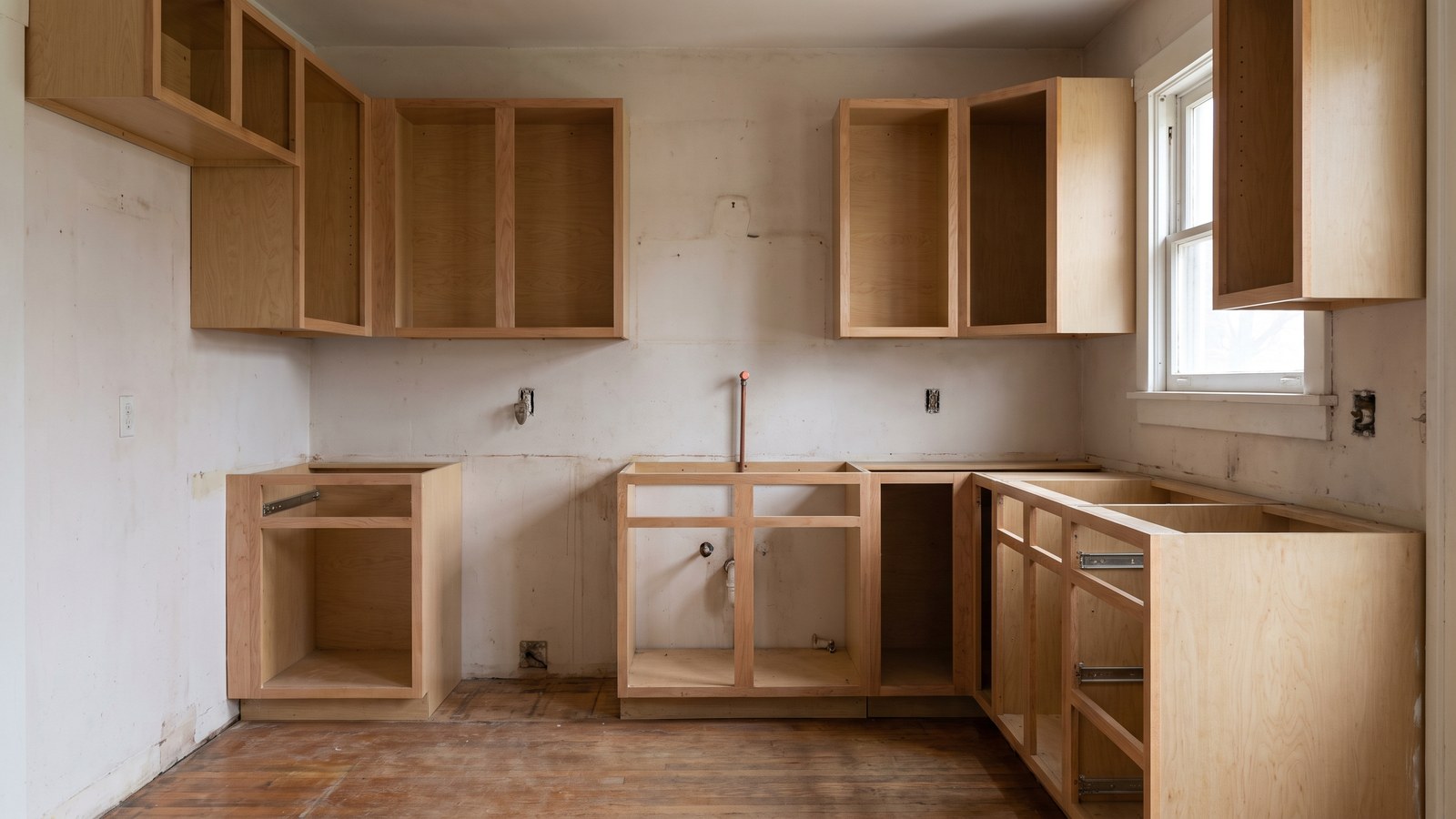 An unfinished kitchen in an older house with cabinet carcasses lacking doors, a gap where an appliance should sit and a capped water line at the wall